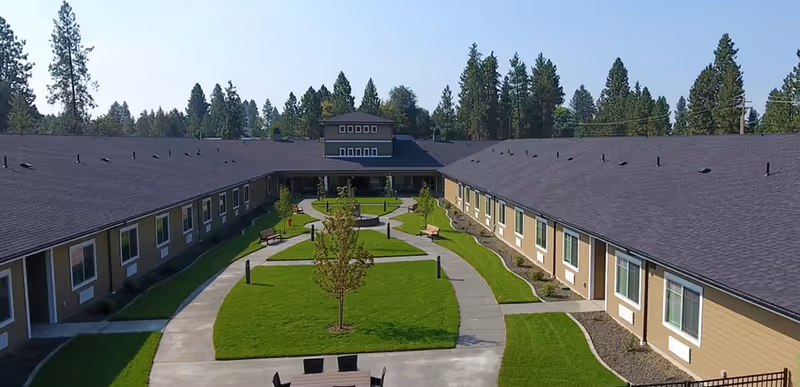 Aerial view of a senior living facility courtyard with a U-shaped building surrounding a green lawn with trees, benches, and paved walkways. The building has multiple windows and a dark roof, with tall pine trees in the background under a clear sky.