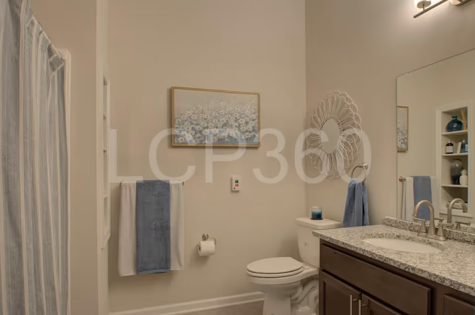 A clean and well-lit bathroom featuring a toilet, a granite countertop with a sink and faucet, a large mirror, a decorative wall mirror, a towel rack with blue and white towels, a shower with a striped curtain, and a framed floral artwork on the wall.