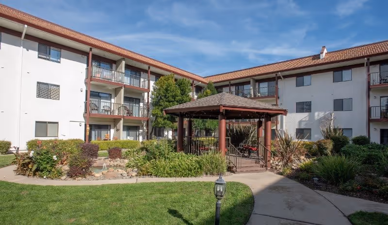 Outdoor courtyard area of a senior living facility with a gazebo surrounded by greenery, shrubs, and a small pond. The building has three stories with balconies and windows overlooking the courtyard under a clear blue sky.