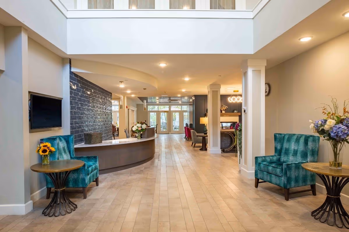 A spacious and well-lit interior lobby area of a senior living facility with a curved reception desk, two teal armchairs each next to a small round table with flower arrangements, light wood flooring, and large windows at the far end letting in natural light.