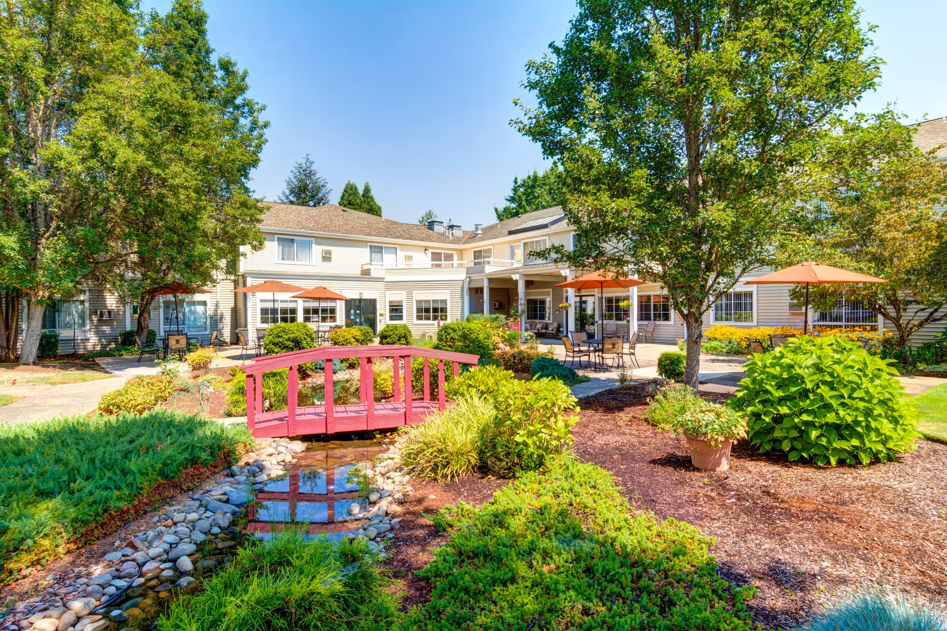 A bright outdoor garden area at The Oaks at Lebanon featuring a small red wooden bridge over a narrow stream, surrounded by lush green bushes, trees, and plants. In the background, there is a two-story building with beige siding, large windows, and several patio tables with orange umbrellas.