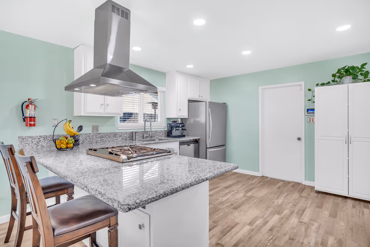 A modern kitchen with light green walls, white cabinets, and a granite countertop island with a built-in gas stove. Two wooden bar stools with dark brown seats are positioned at the island. Stainless steel refrigerator and dishwasher are visible, along with a coffee machine on the counter. A fire extinguisher is mounted on the wall near the window with white blinds. A white door and a white cabinet with a green plant on top are on the right side of the room. The floor is light wood, and recessed ceiling lights illuminate the space.
