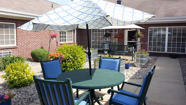 Outdoor patio area with a round green table and four blue cushioned chairs under a large white umbrella with a blue checkered pattern. The patio is surrounded by a garden with green bushes, flowers, and gravel. In the background, there is a brick building with windows and a door leading inside.
