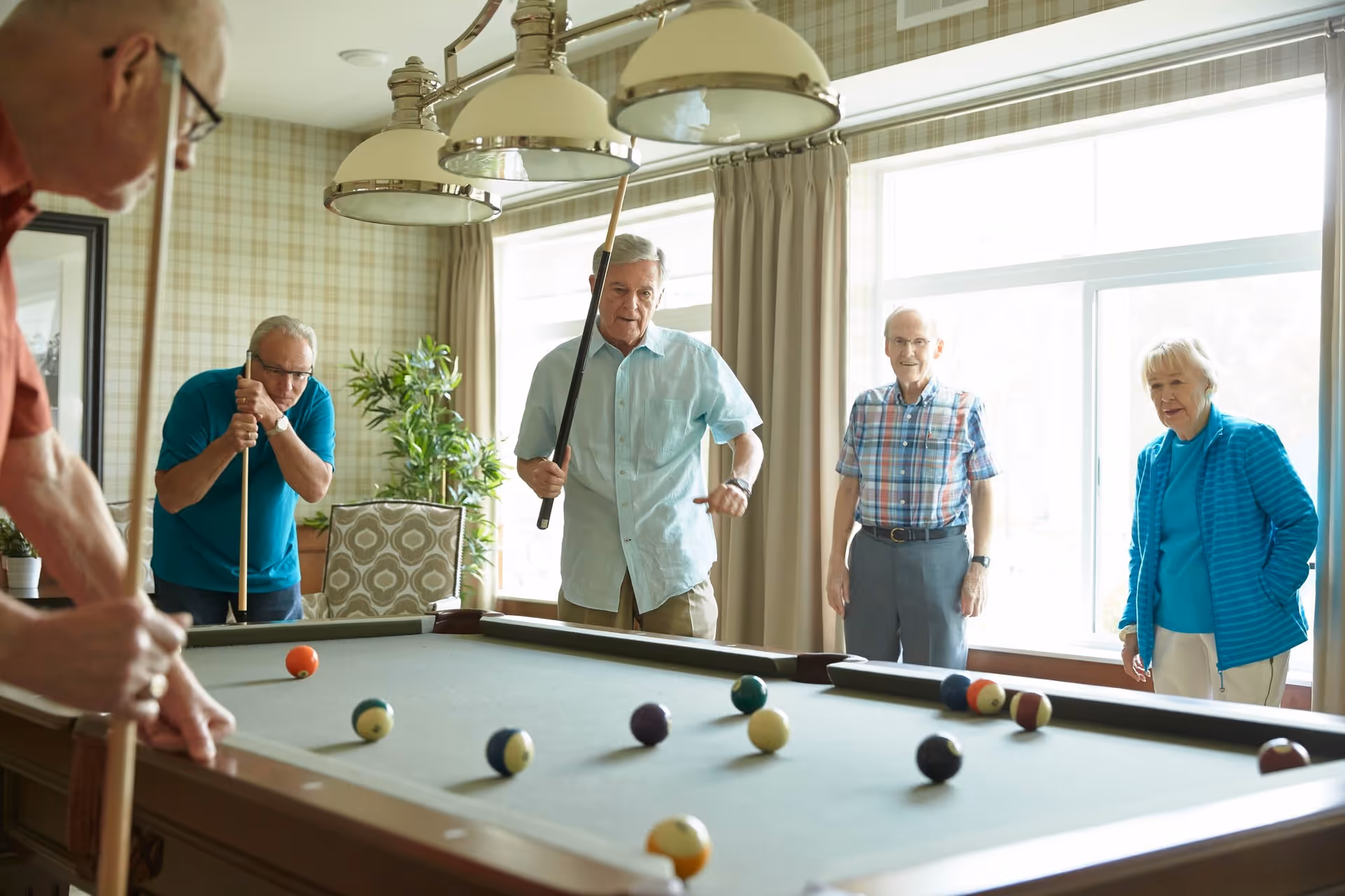 A group of four elderly people playing pool in a well-lit room with large windows and patterned wallpaper. Two men are holding pool cues and appear to be taking shots, while another man and a woman watch. The room has a cozy and social atmosphere.
