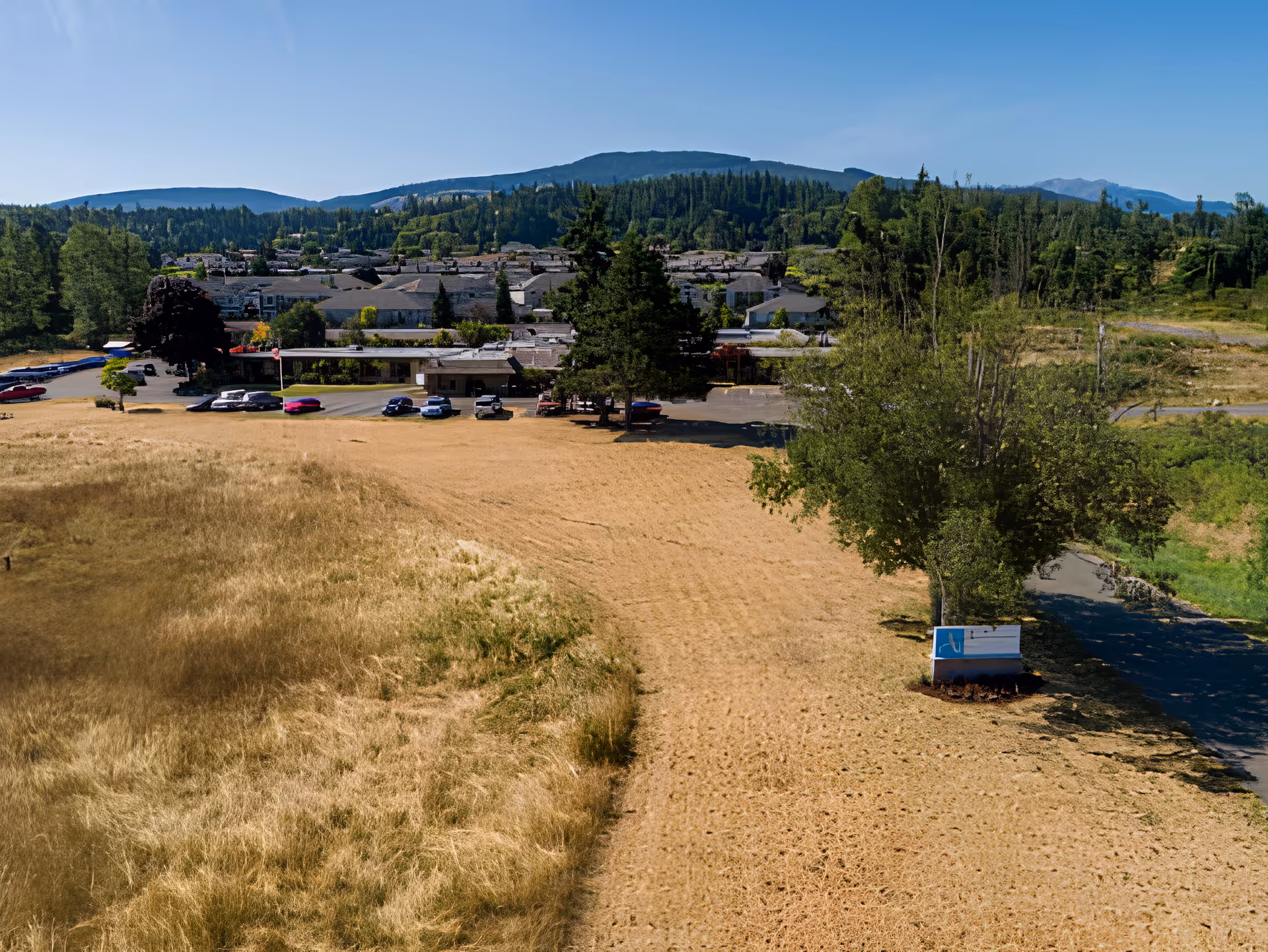 Aerial view of Avamere Olympic Rehabilitation of Sequim facility surrounded by dry grassy fields and trees, with mountains in the background under a clear blue sky. Several cars are parked in front of the building.