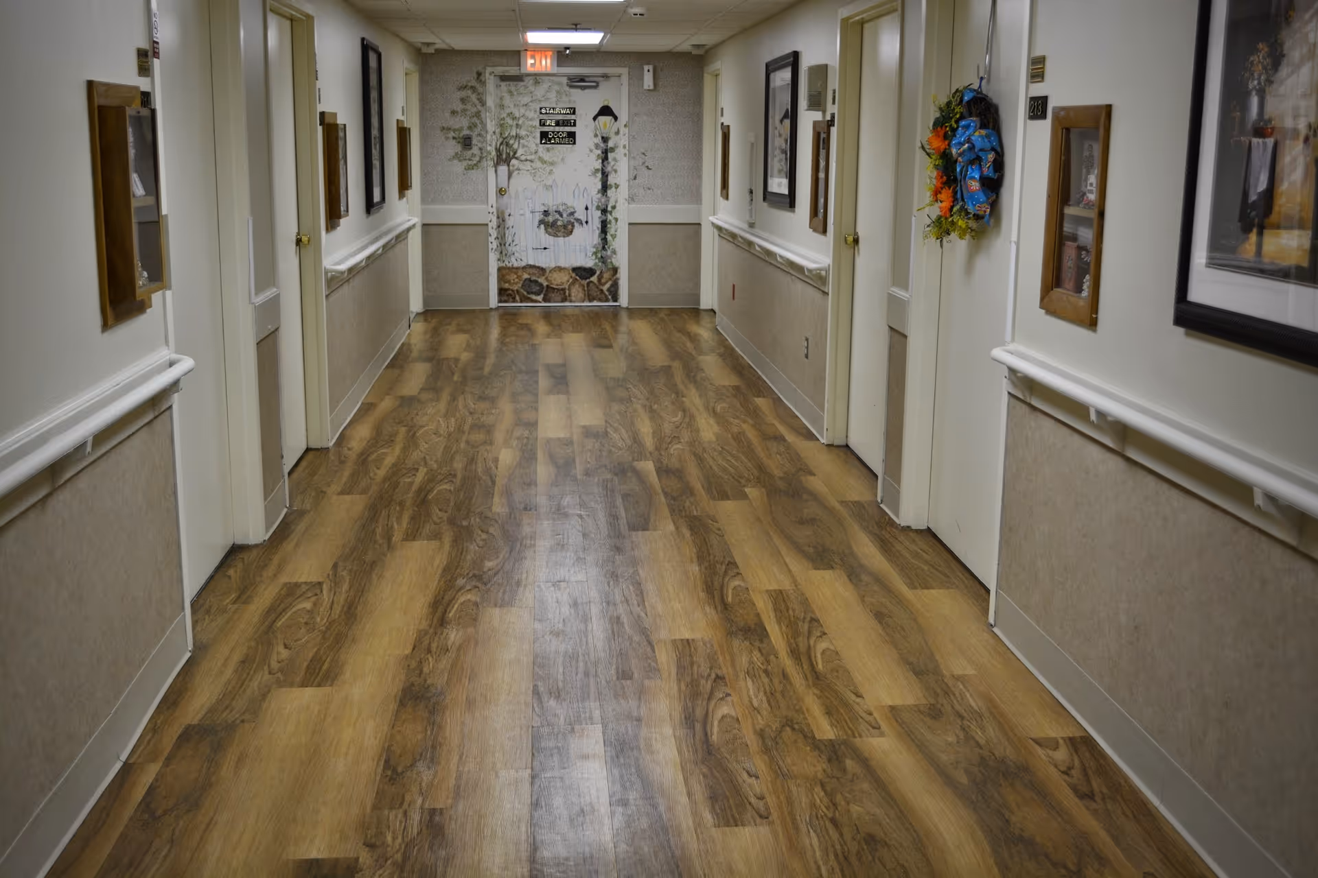 A clean, well-lit hallway in a senior living facility with wood-patterned flooring, white walls, handrails on both sides, several closed doors, framed pictures on the walls, and a decorated door at the end of the hallway.