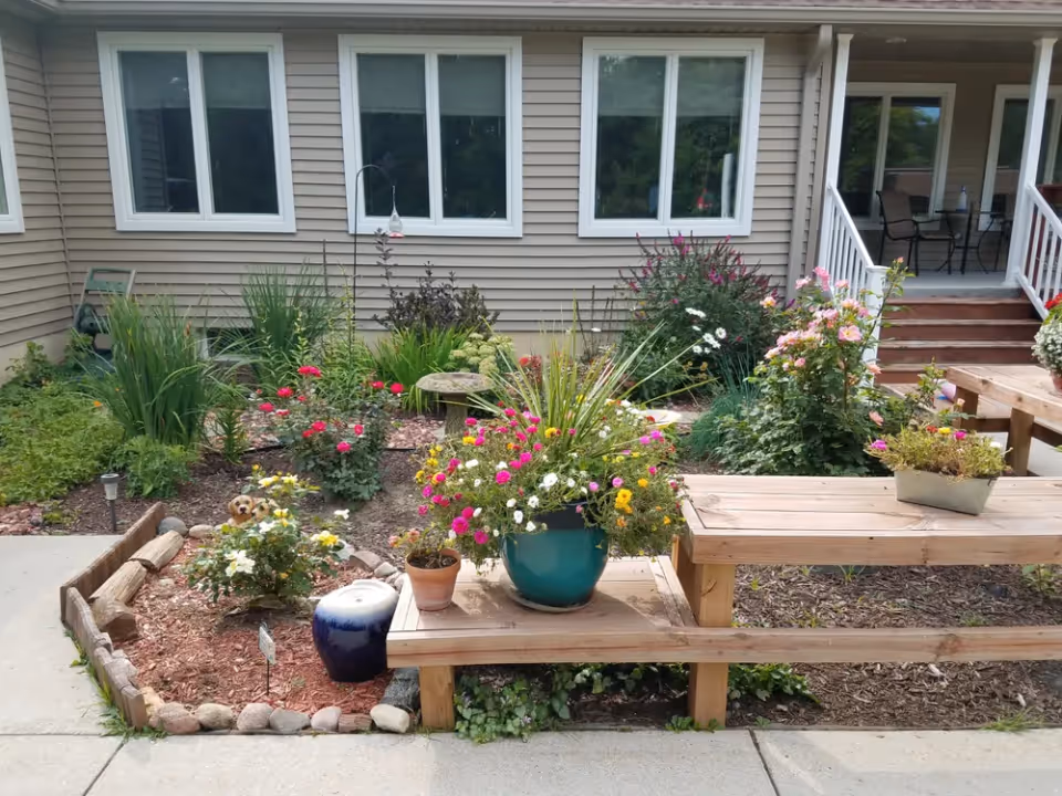 A garden area with various colorful flowers and plants in pots and flower beds in front of a beige building with white-framed windows. There are wooden benches and a small bird feeder hanging near the windows. A porch with chairs is visible on the right side.