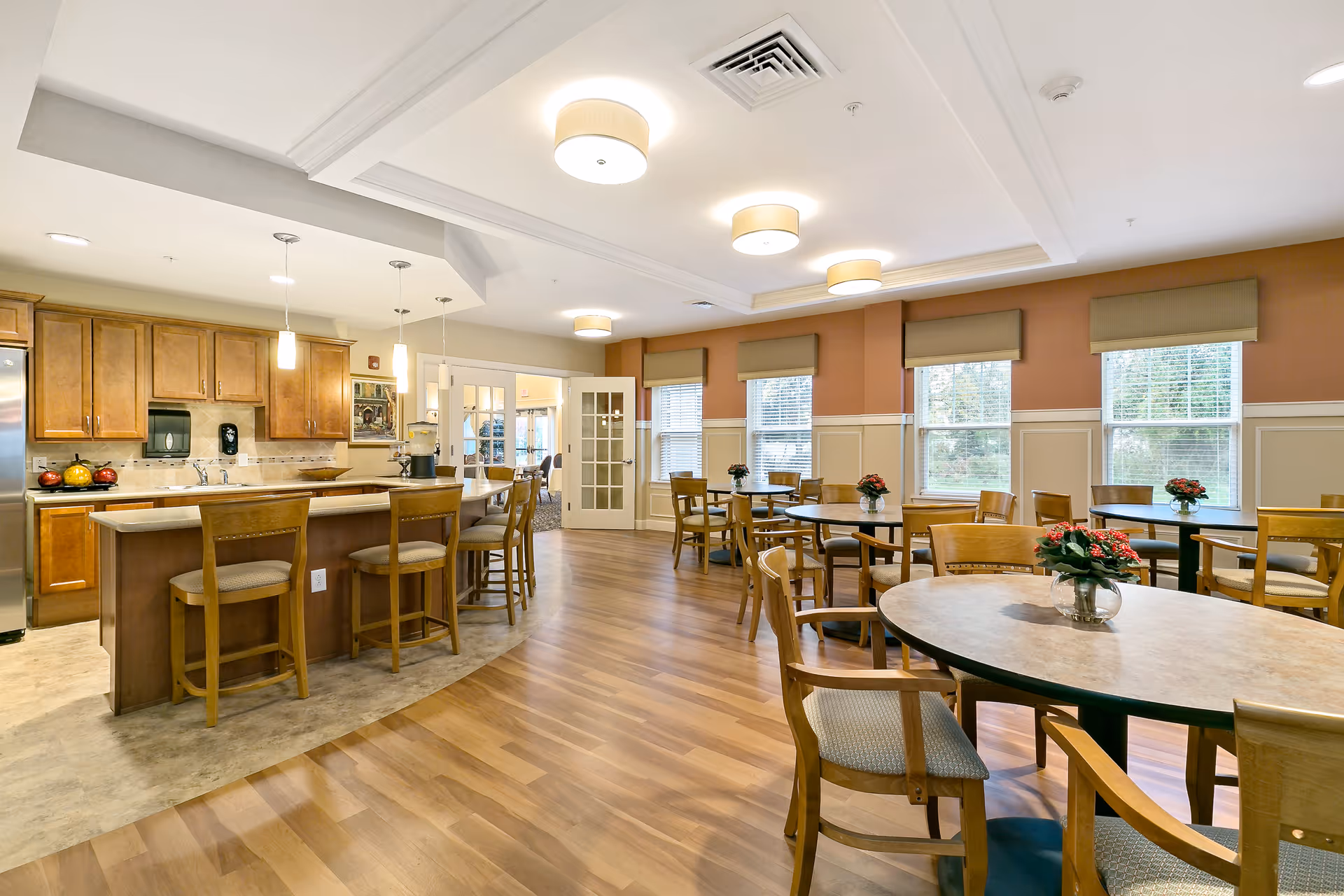 A spacious dining area in an assisted living facility featuring several round tables with wooden chairs, each table decorated with a small flower arrangement. The room has large windows with blinds, warm-colored walls with white wainscoting, and a kitchen area with wooden cabinets, a countertop with bar stools, and pendant lighting.