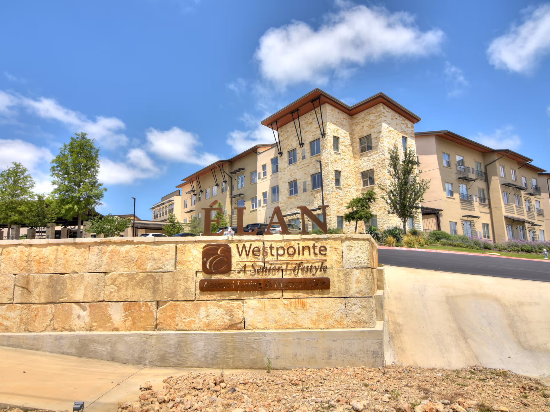 Exterior view of Élan Westpointe at New Braunfels, a senior living facility with a multi-story building made of stone and beige stucco. The facility name is displayed on a stone wall in front, with a clear blue sky and some trees in the background.
