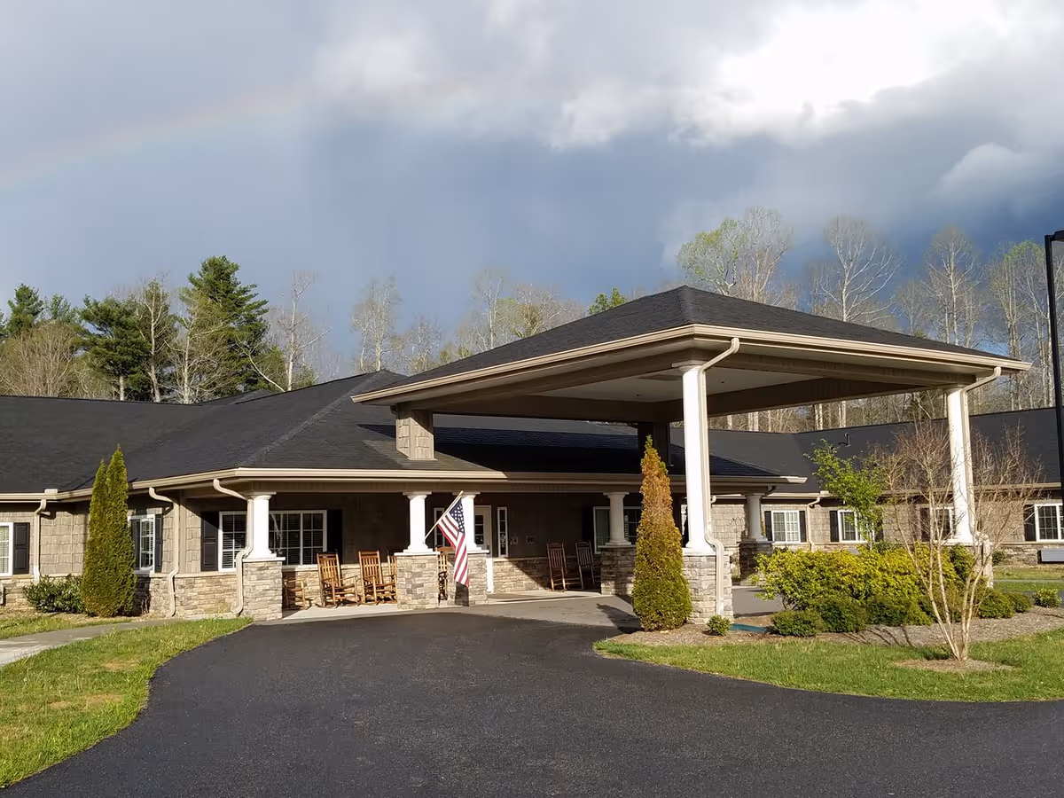 Front entrance of a single-story senior living building with a covered porte-cochere, rocking chairs, an American flag, and landscaping under a cloudy sky.