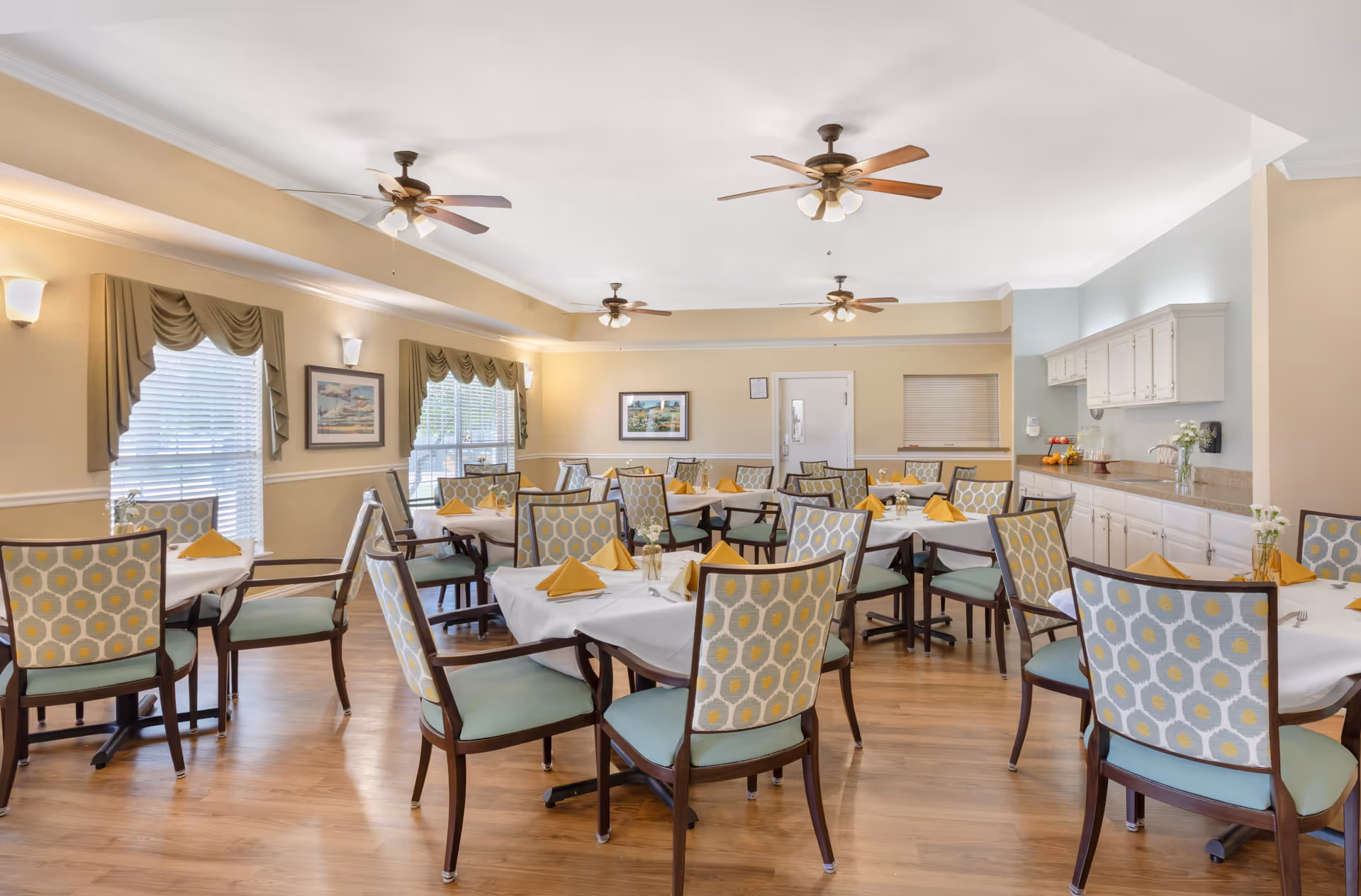 A bright dining room with multiple tables covered in white tablecloths and set with yellow folded napkins. The chairs have patterned backs with blue and yellow designs. The room has wooden flooring, beige walls, three ceiling fans with lights, and windows with green valances. There are framed pictures on the walls and a counter with cabinets on the right side.