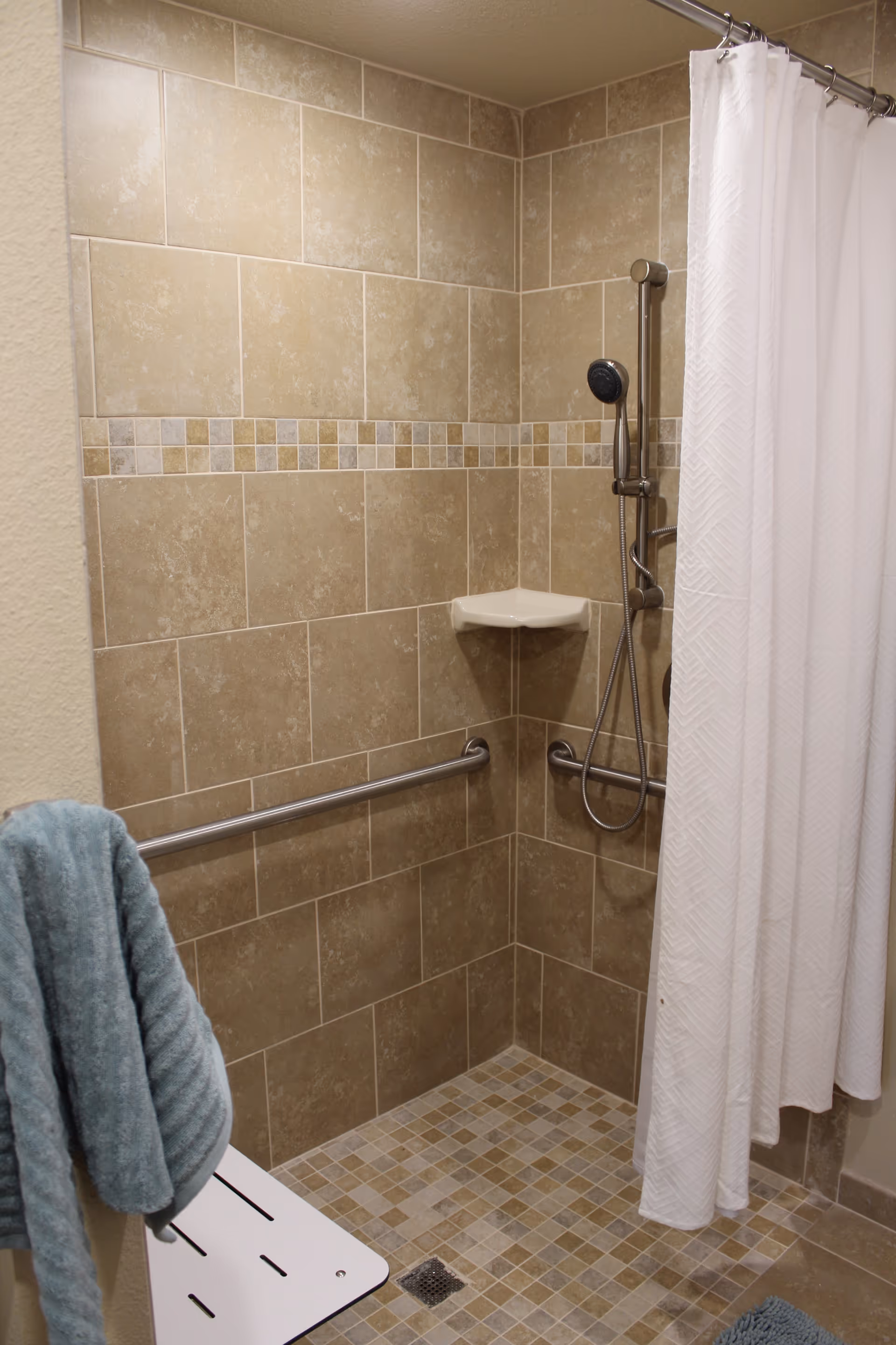 A tiled shower area with beige and brown tiles, a white shower curtain, a handheld showerhead mounted on a vertical bar, a corner soap dish, a horizontal grab bar, and a white fold-down shower seat with a blue towel hanging on it.