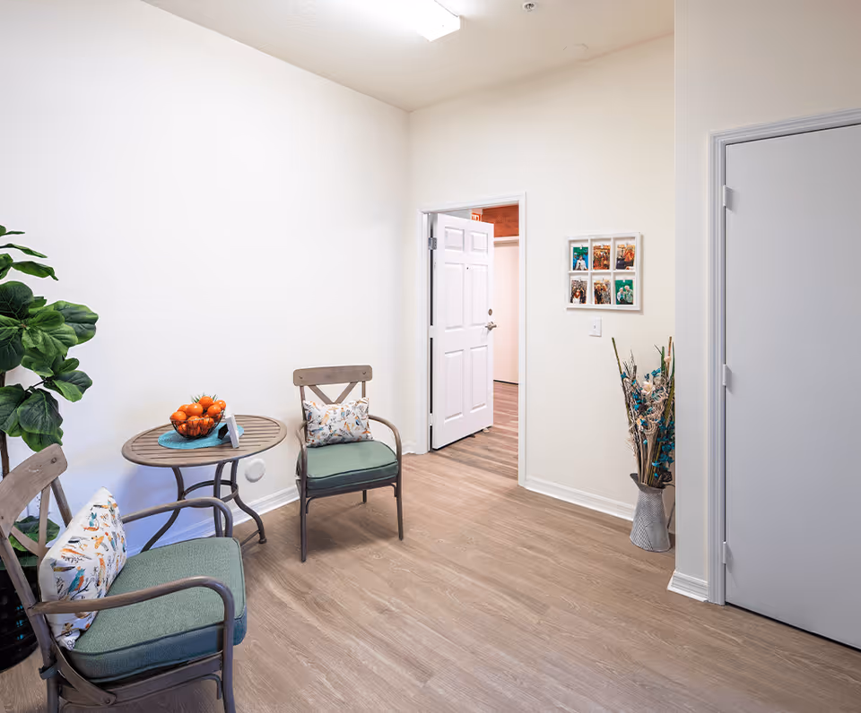 A small seating area with two wooden chairs with green cushions and decorative pillows, a round wooden table with a bowl of oranges, a potted plant, a white door slightly open leading to another room, a wall with framed photos, and a vase with dried flowers on a wooden floor.