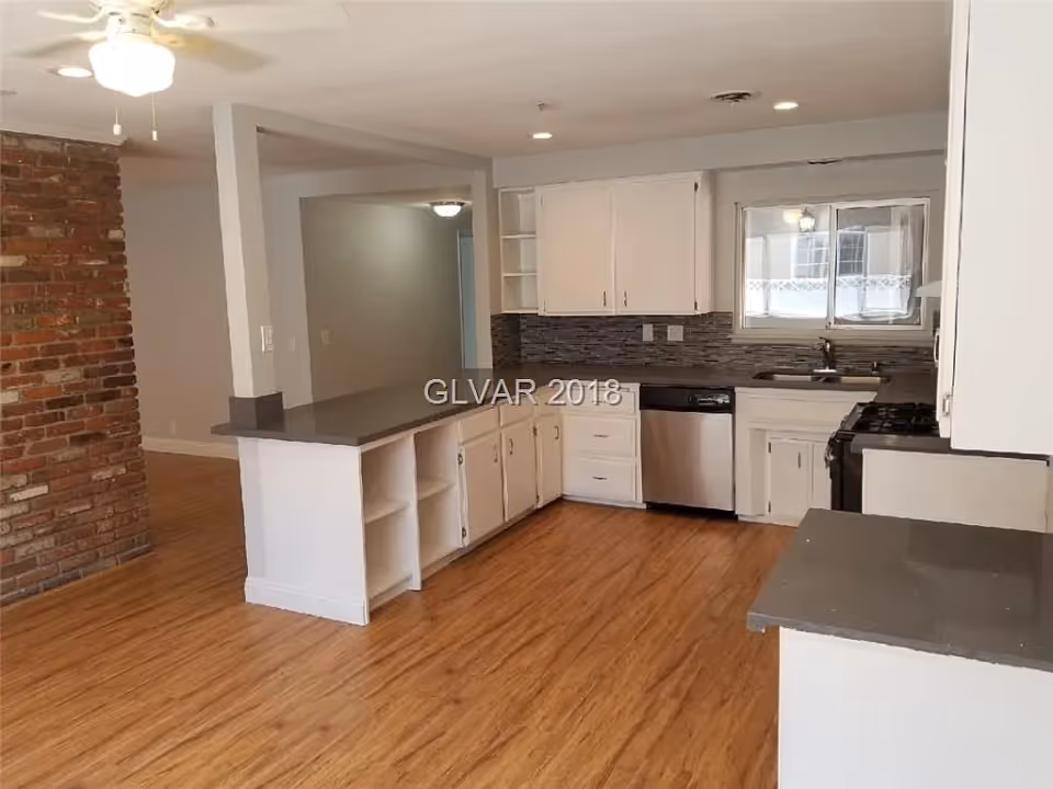 Interior view of a kitchen with white cabinets, gray countertops, a stainless steel dishwasher, a gas stove, a window above the sink, and a ceiling fan. The kitchen has wood flooring and an exposed brick wall on the left side.