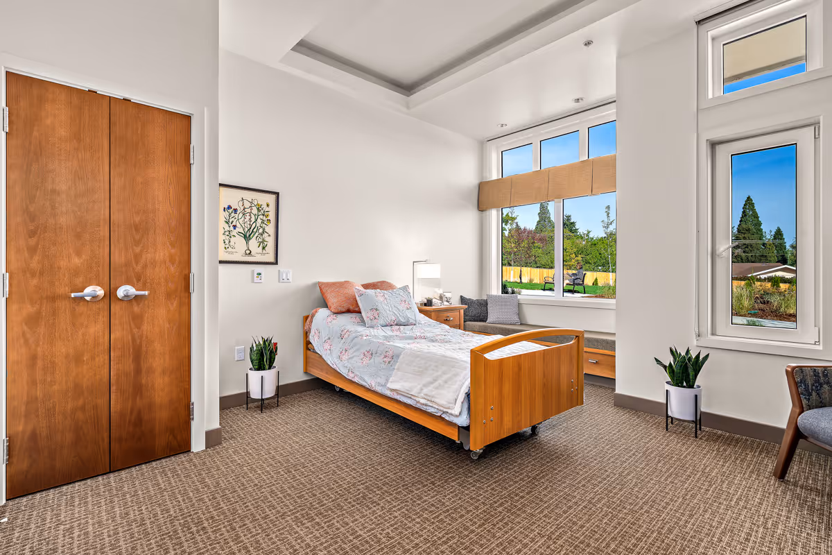 A bright and spacious bedroom in a senior living facility with a single wooden bed featuring floral bedding and pillows. The room has large windows with a view of greenery outside, a wooden nightstand with a lamp, a cushioned window seat with pillows, two potted plants, a wooden double door, and a framed botanical print on the wall.