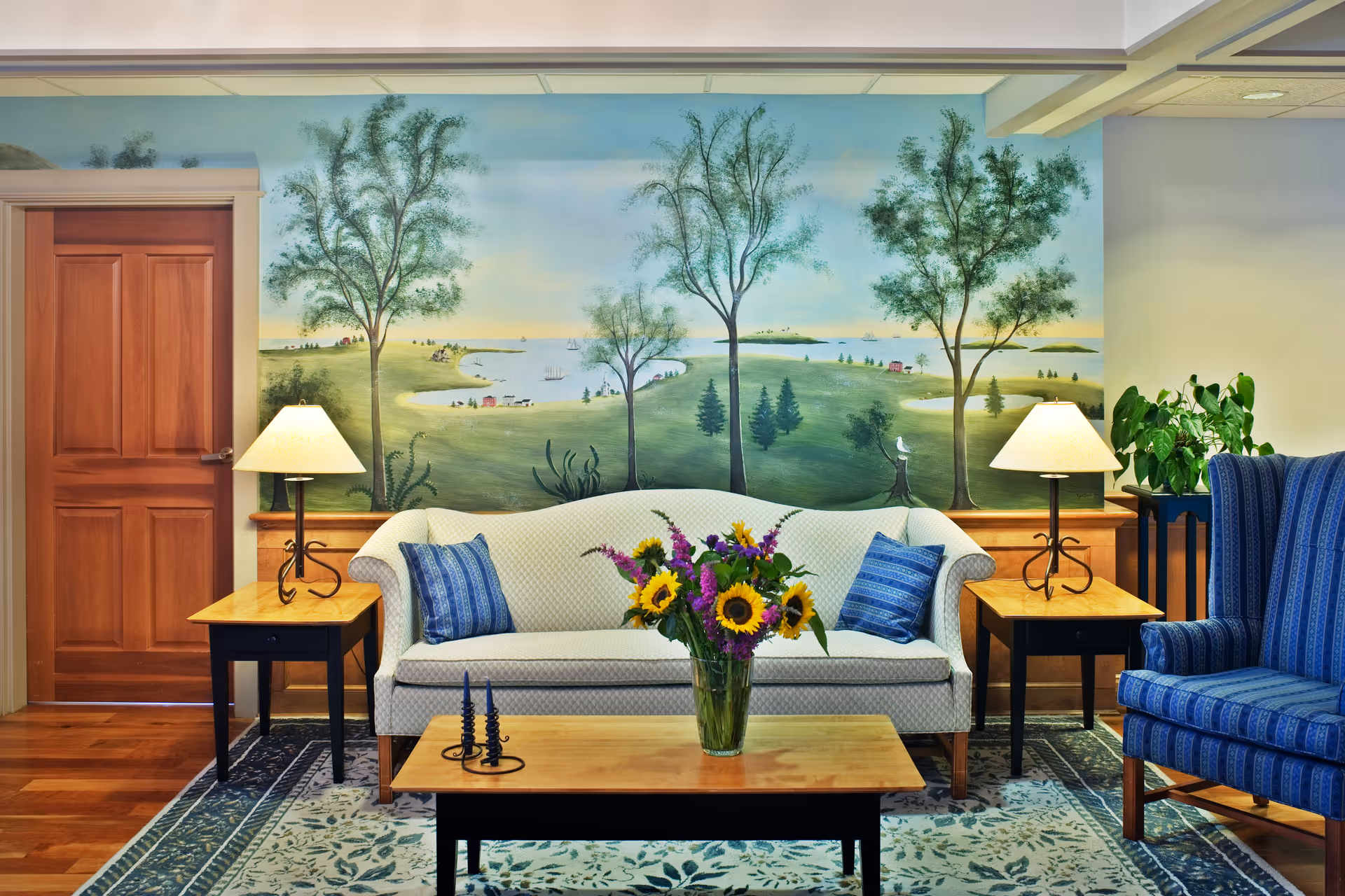 A cozy living room with a white sofa adorned with two blue patterned pillows, a wooden coffee table with a vase of sunflowers and other flowers, two wooden side tables each with a lamp, a blue striped armchair, a leafy green plant on a small table, and a large mural on the wall depicting a scenic landscape with trees, water, and small houses.
