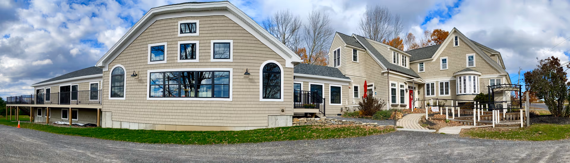 Exterior panoramic view of a large, beige senior living facility building with multiple windows, a paved walkway, and some landscaping including small trees and bushes under a partly cloudy sky.