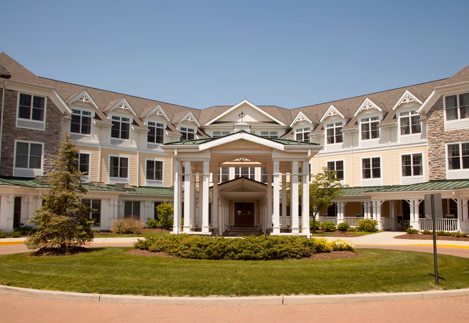 Front entrance of a multi-story senior living building with a covered portico, landscaped roundabout, and many windows.