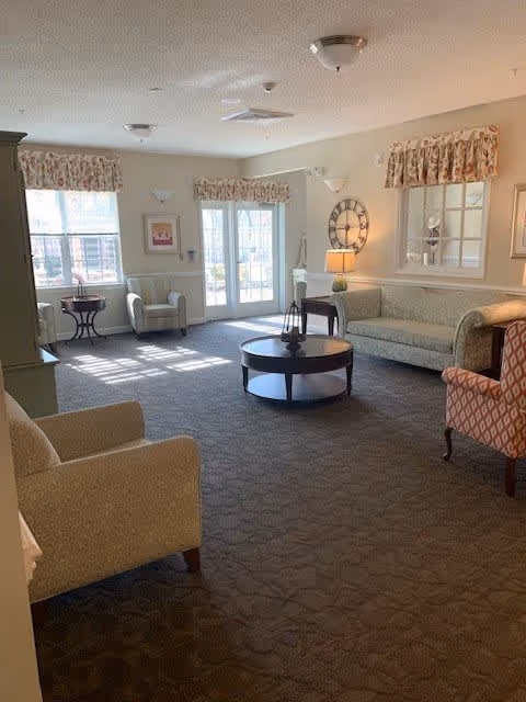 A well-lit living room with beige walls and carpeted floor. The room features a light green sofa, two armchairs with patterned upholstery, a round wooden coffee table with a decorative lantern, and a small side table with a lamp. There are two large windows and a glass door with floral valances, allowing natural light to fill the space. A large decorative wall clock is mounted on the wall above the sofa.