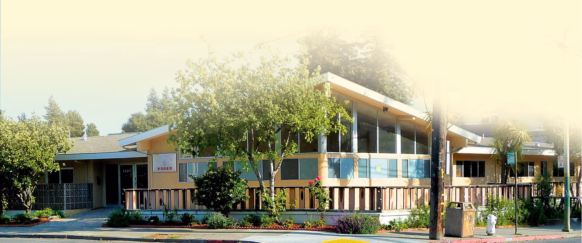Exterior view of a single-story assisted living facility building with large windows, surrounded by trees and landscaping, located on a street corner with a sidewalk and street signs.