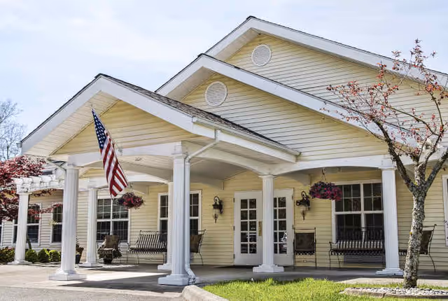 Exterior view of a single-story assisted living facility with a covered porch supported by white columns. The building is painted light yellow with white trim. There are hanging flower baskets, benches on the porch, an American flag, and a small tree with red leaves in front.