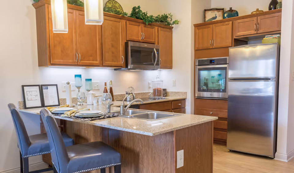 A bright modern kitchen featuring a granite-topped island with sink and bar stools, wooden cabinets and stainless steel appliances.