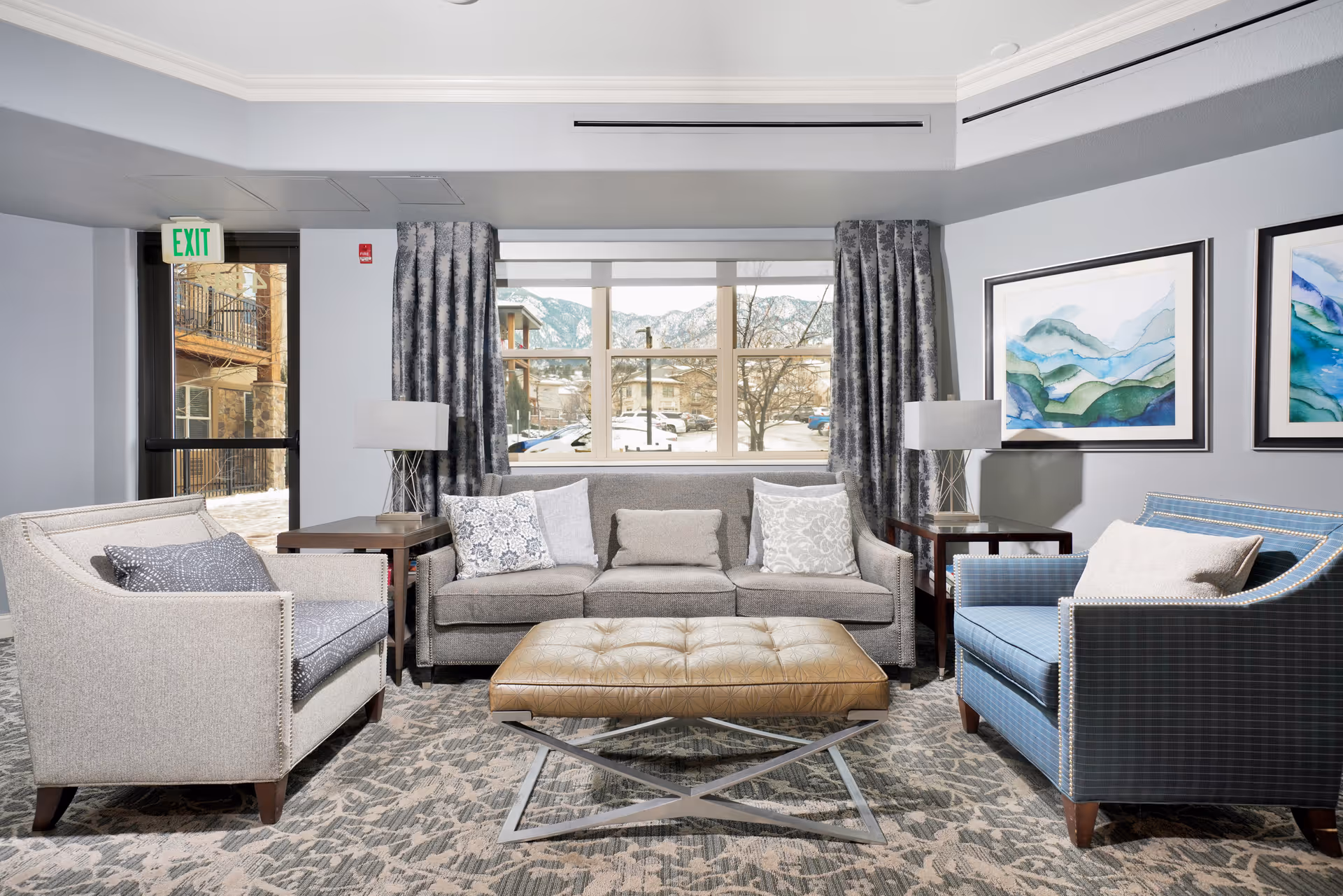 A bright and modern living room with a large window showing an outdoor view of buildings and mountains. The room features a gray sofa with multiple cushions, two armchairs—one beige and one blue with a pillow—and a large tufted leather ottoman in the center. There are two side tables with matching lamps, patterned curtains, and two framed abstract landscape paintings on the wall.