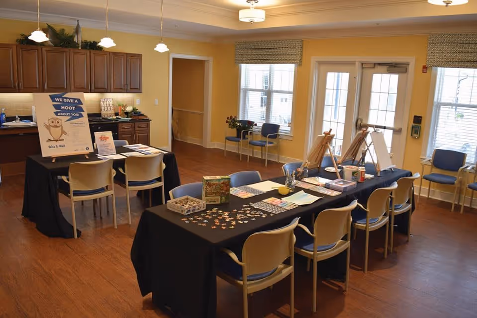 A well-lit activity room with yellow walls and wooden flooring. There are two tables covered with black tablecloths; one table has chairs around it and is set up with art supplies including easels, paintbrushes, and puzzle pieces. The other table has informational signs and pamphlets. The room has large windows with blinds and a glass door letting in natural light. Blue and beige chairs are arranged around the room, and wooden cabinets are mounted on the wall in the background.