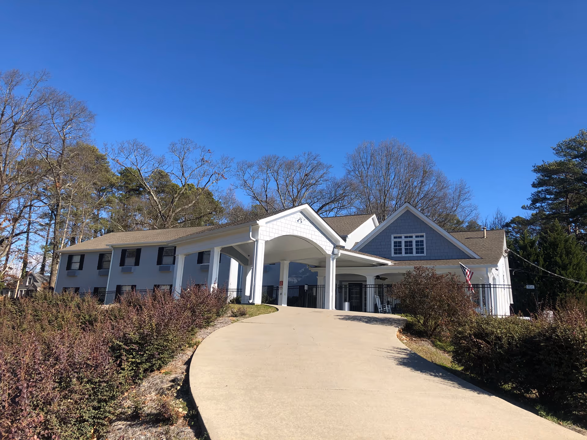 Front exterior of a two-story senior living building with a covered entrance, curved driveway, and surrounding landscaping under a clear blue sky.