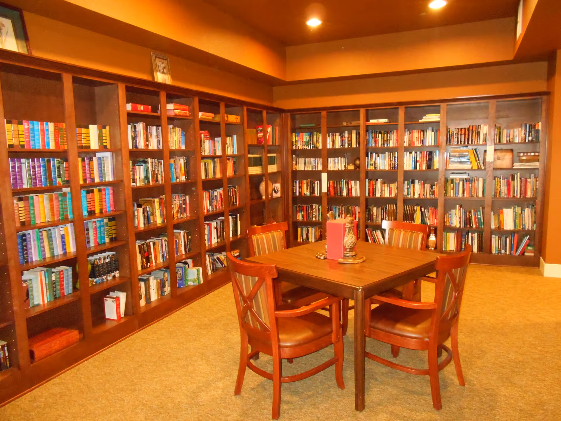 A cozy library room with wooden bookshelves filled with books lining the walls. In the center, there is a wooden table with four wooden chairs that have cushioned seats and backs. The room has warm lighting and a carpeted floor.