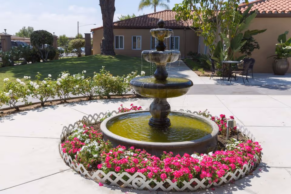 Outdoor courtyard area with a three-tiered water fountain surrounded by a circular flower bed with pink and white flowers. In the background, there is a building with a tiled roof, some greenery, and a small table with chairs on a paved surface.