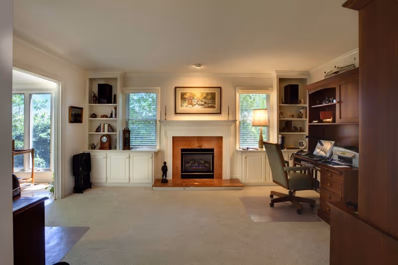A cozy living room with a fireplace centered between two windows with white blinds. Built-in white shelves and cabinets flank the fireplace, displaying decorative items and books. To the right, there is a wooden desk with a computer and office chair, illuminated by a floor lamp. The room has beige carpeting and light-colored walls, with natural light coming in from a doorway on the left.