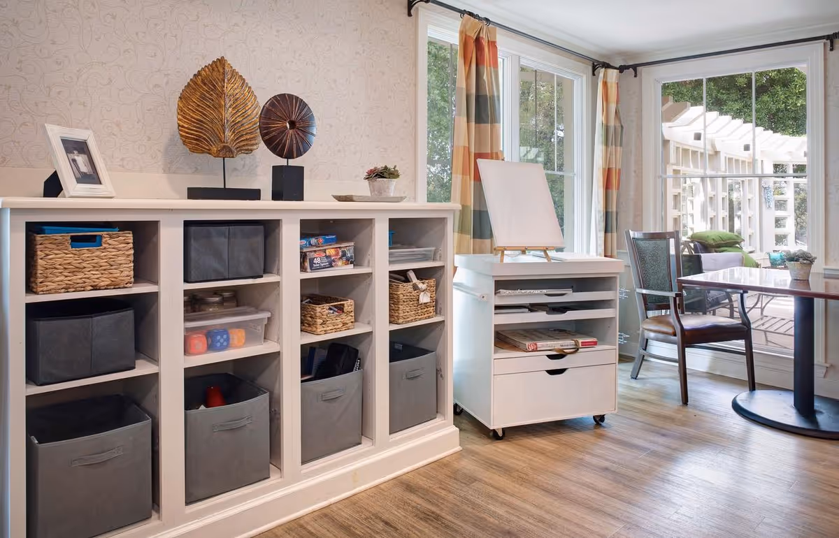 A bright room with a white storage cabinet filled with baskets and boxes, a small white rolling cart with an easel on top, a wooden chair, and a table near large windows with colorful plaid curtains. Outside the windows, there is a patio area with seating and greenery.