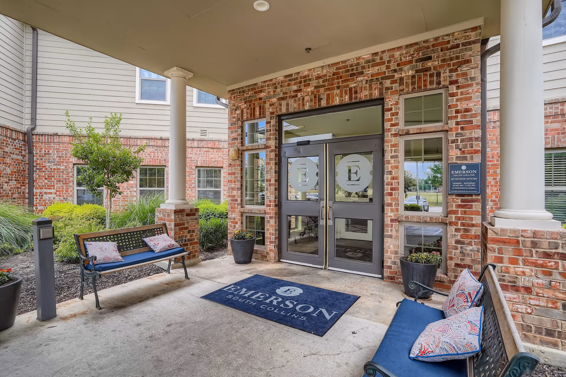 Covered entrance area of Emerson South Collins facility with two benches featuring blue cushions and patterned pillows, potted plants, brick walls, and double glass doors with the Emerson logo. A blue mat with the Emerson South Collins name is placed on the ground in front of the doors.