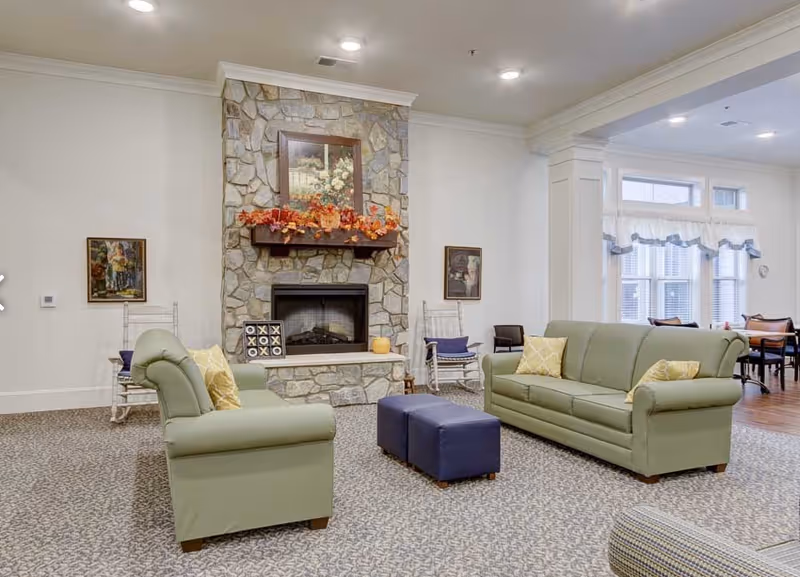 A cozy senior living common area featuring two green sofas with yellow pillows, a stone fireplace decorated with autumn leaves and a framed painting above it, two white rocking chairs, and a pair of blue ottomans. The room has carpeted flooring and large windows with white curtains allowing natural light.