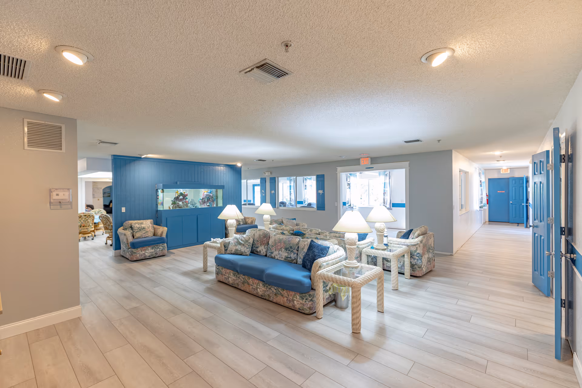A bright and spacious common area in an assisted living facility featuring floral patterned sofas and armchairs with blue cushions, white wicker side tables with lamps, light wood flooring, and a large built-in aquarium set into a blue accent wall. The room has multiple windows and blue doors along a hallway.