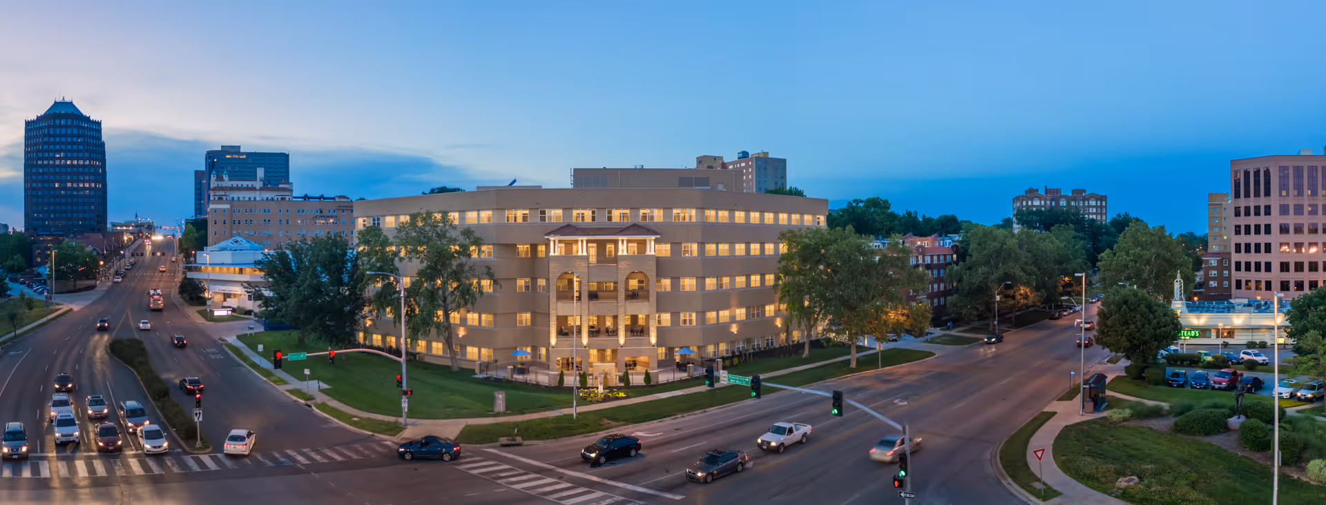 Panoramic evening view of a multi-story senior living building lit up at a busy city intersection.