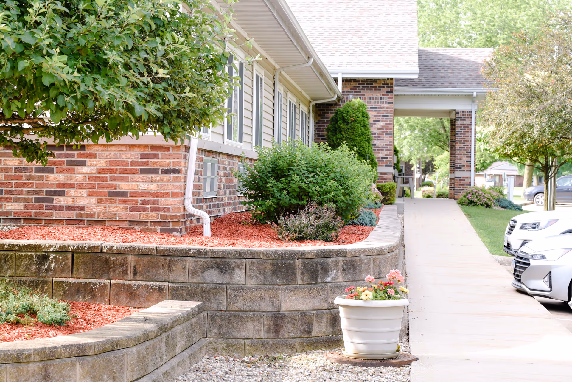 Exterior view of a senior living facility with a brick and siding building, a landscaped garden bed with bushes and mulch, a white flower pot with colorful flowers, a concrete walkway leading to a covered entrance, and parked cars along the side.