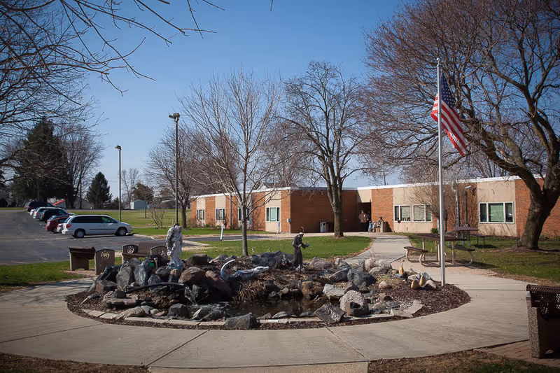 Outdoor view of a single-story brick building with a small pond and rock garden in the foreground, surrounded by a concrete walkway. There are leafless trees, a flagpole with an American flag, picnic tables, and parked cars visible on the left side under a clear blue sky.
