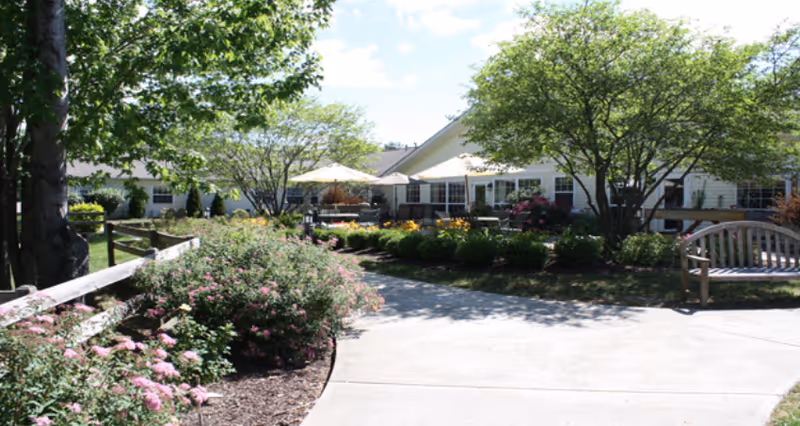 Outdoor garden area with a concrete pathway, wooden bench, flowering bushes, and trees in front of a single-story building with patio umbrellas and seating.