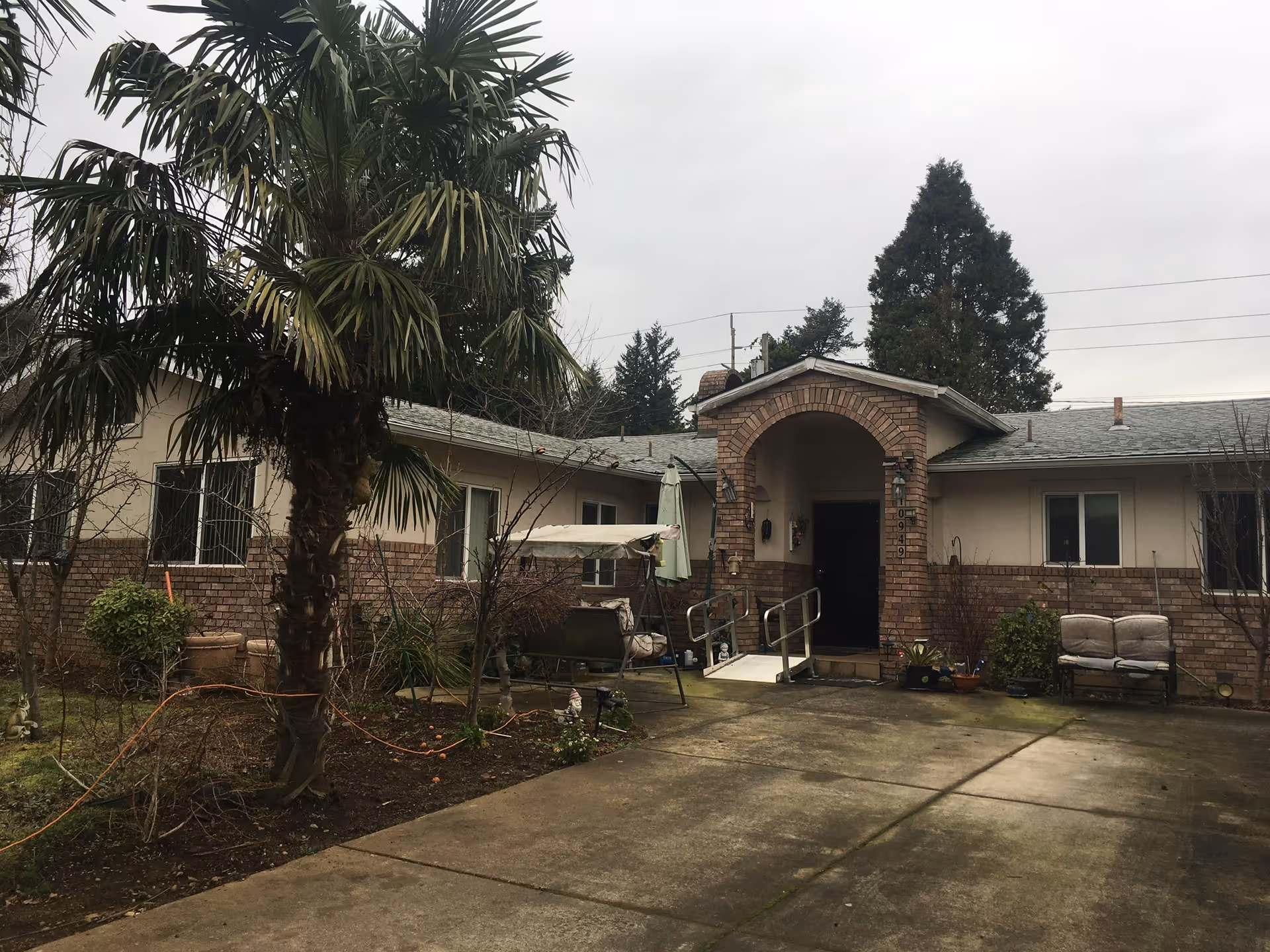 Exterior view of a single-story building with a brick and beige facade, featuring an arched entrance with a ramp. There are palm trees and other plants in the garden area, a swing bench, and outdoor seating. The sky is overcast.