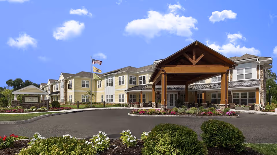 Front exterior view of a large two-story assisted living nursing home building with a covered entrance supported by wooden beams. The building is painted yellow with white trim and has multiple windows. There are landscaped flower beds and bushes around the driveway, and two flagpoles with flags flying in front of the building under a blue sky with scattered clouds.