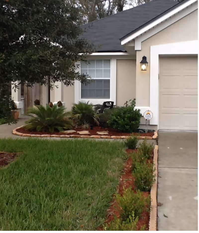 Front yard of a house with a well-maintained lawn, landscaped garden beds with shrubs and small palm plants, a concrete driveway leading to a closed garage door, and a window with white trim. A porch light is on near the garage.