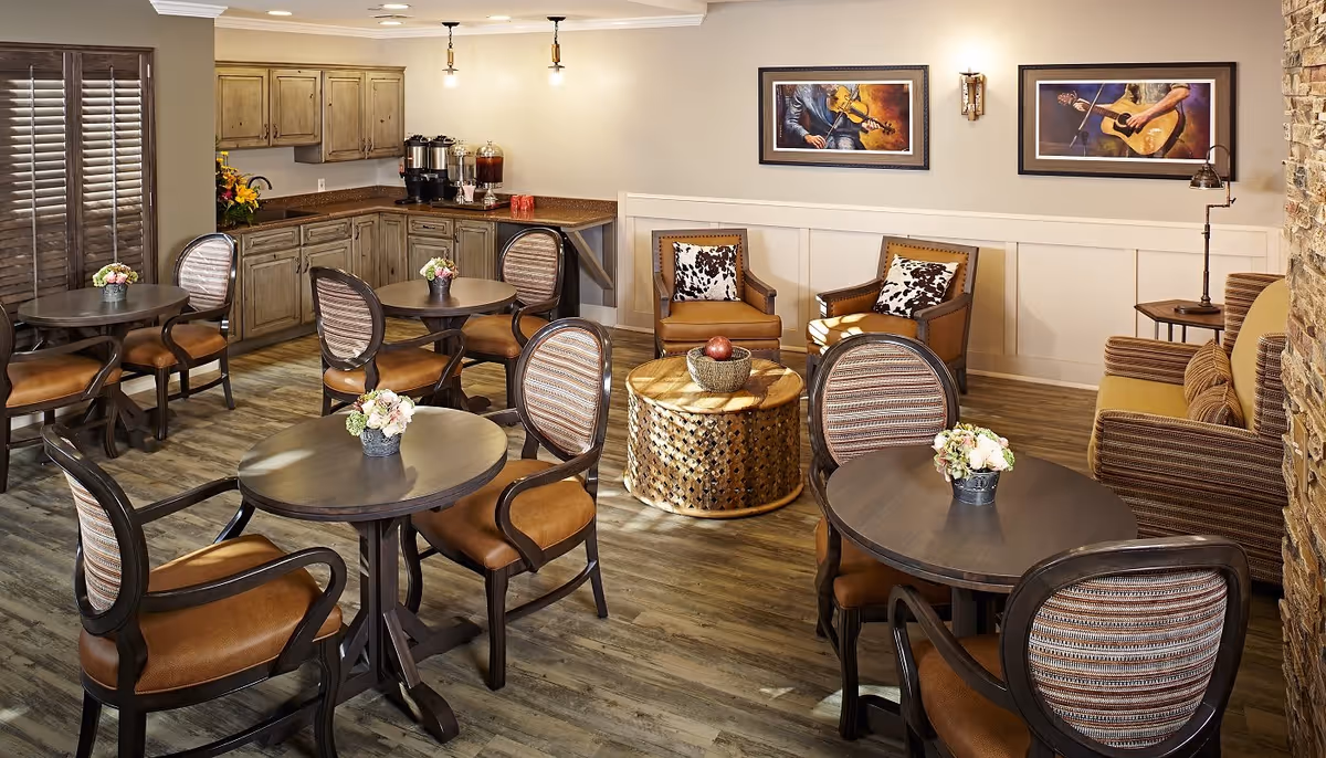 A cozy senior living common area with several round tables and cushioned chairs arranged around the room. There is a small kitchenette with cabinets, a coffee and beverage station, and a countertop in the back corner. The room features wood flooring, warm lighting, and framed artwork on the walls. Comfortable armchairs and a sofa are placed near a round wooden coffee table with decorative items on it.