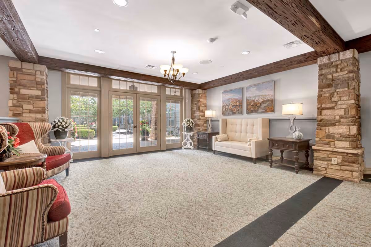 A bright and spacious living room area with large glass doors leading to an outdoor garden. The room features stone pillars, wooden ceiling beams, a beige tufted sofa, two side tables with lamps, two floral paintings on the wall, and two striped armchairs with red cushions. There are also flower arrangements on small tables near the seating.