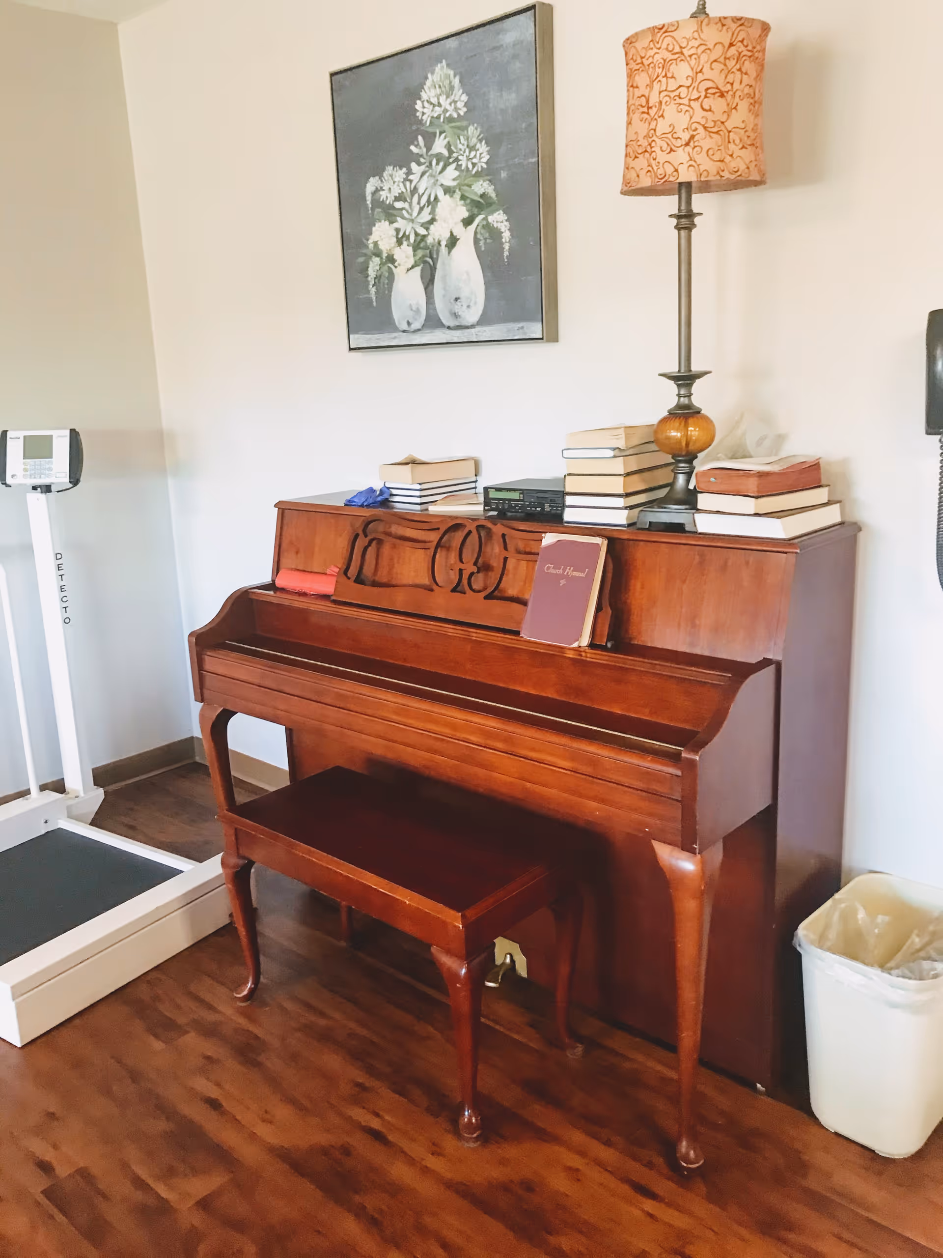 A wooden upright piano with a matching bench in front of it, topped with a stack of books, a lamp with an orange patterned shade, and a small electronic device. Above the piano hangs a painting of white flowers in vases. To the left of the piano is a white medical scale, and to the right is a white trash bin on a wooden floor.