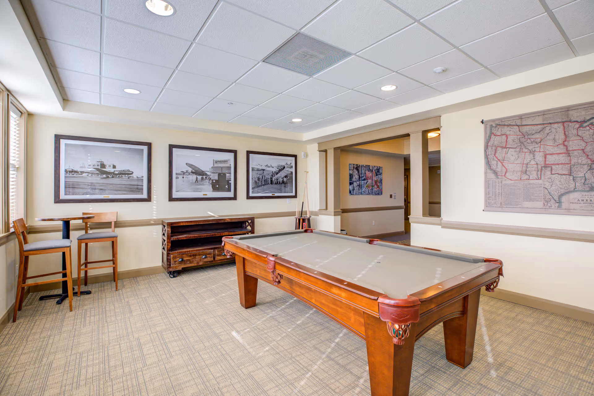 Bright communal game room featuring a pool table, bar-height seating, framed photos on the wall, and a large map.