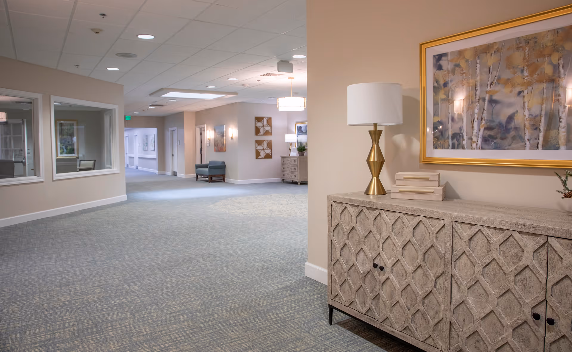 A well-lit interior hallway of a senior living facility with beige walls and carpeted floors. On the right side, there is a decorative wooden cabinet with a gold and white lamp, two stacked books, and a framed abstract painting above it. The hallway extends into the distance with seating areas and wall art visible along the corridor.