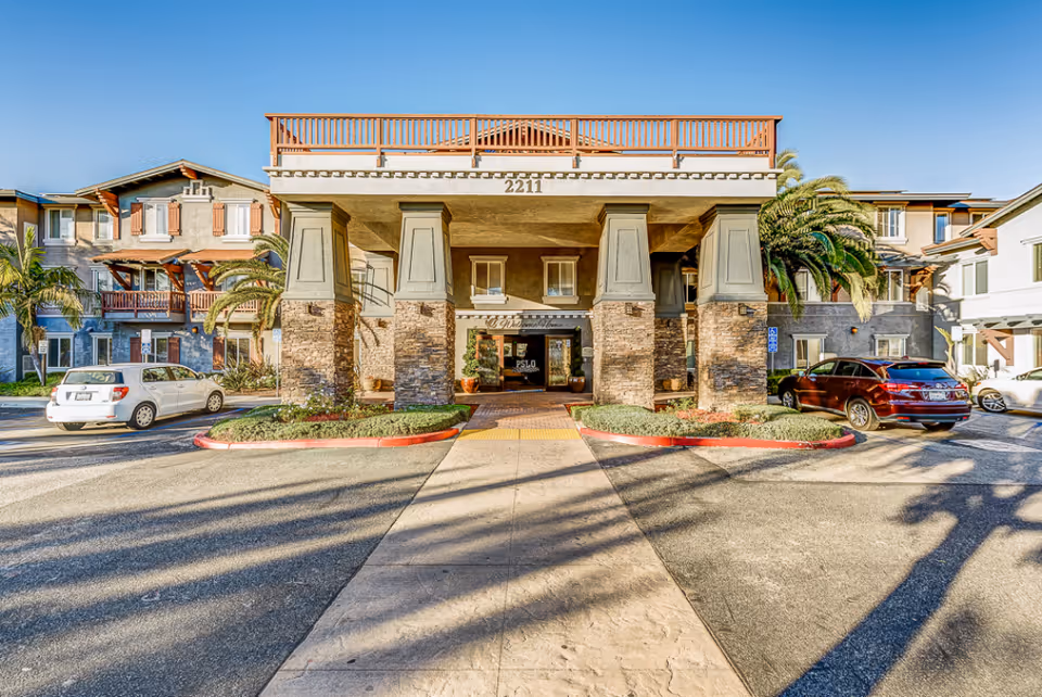 Front entrance of a multi-story senior living building with a covered porte-cochère, stone columns, landscaping, and parked cars.