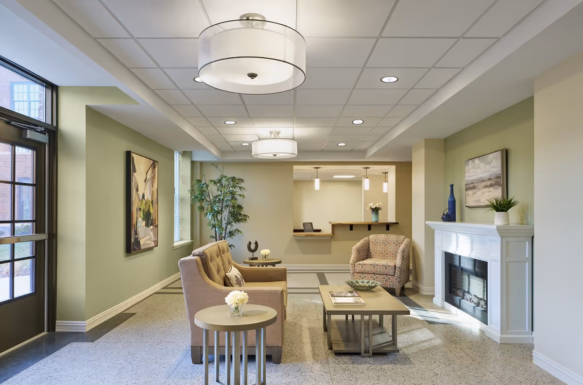 A cozy living room area in Bardwell Residences featuring a beige sofa, a patterned armchair, a wooden coffee table with magazines, and a side table with a small flower vase. The room has light green walls, a white fireplace with decorative items on the mantel, a large window, and modern ceiling lights.